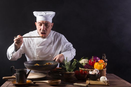 Male chef in white uniform is tasting food by using a wooden ladle at the kitchen of a restaurant before serving while working in a restaurant kitchen