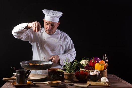 Male cook in white uniform and hat putting pot herbs on cooking pan before serving while working in a restaurant kitchen