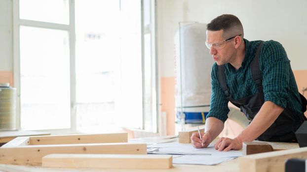 A portrait of a carpenter in goggles and work overalls draws a blueprint for a workshop