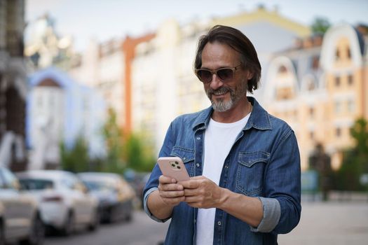 Texting or posting in social media post, photo mature handsome man with smartphone outdoors wearing denim shirt and sunglasses. Handsome middle aged man with grey hair making photos while traveling.