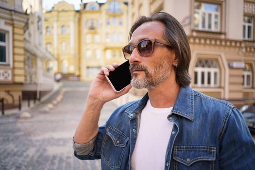 Middle aged man with grey bearded talking on the phone standing outdoors in old city background wearing denim jeans shirt. Mature business man working on the go. Freelancer traveling man.