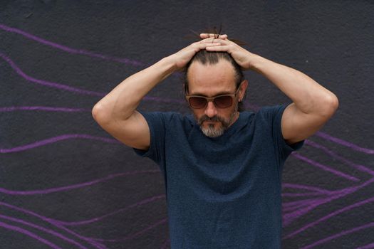 Thoughtful mature man standing with hand folded above his head next to dark urban wall leaned head down wearing sunglasses, dark blue t-shirt and light shorts. Stress, middle age crisis man concept.