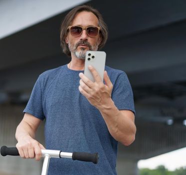 Mature man looking at phone in city. Man made stop using scooter answer the phone. Travel concept. Handsome stylish sexy middle aged grey bearded man in sunglasses under the bridge wearing casual.