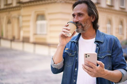 Mature man with coffee and smartphone outdoors. Middle aged man read having video call holding phone and take away paper cup with coffee wearing denim shirt. Business on the go concept.