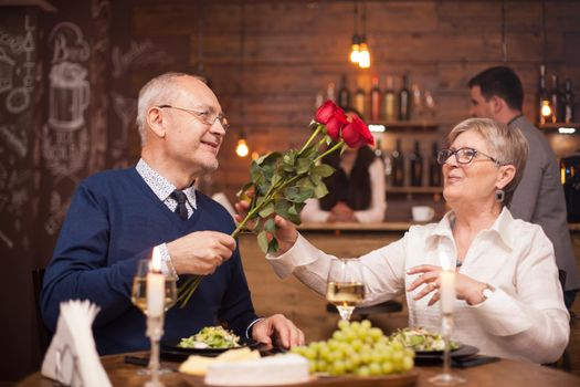 Woman in her sixties happy to recieve roses from her husband during dinner. Senior couple date. Fresh grapes. Cheerful old couple.