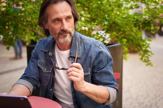 Middle age freelancer businessman working outdoors sitting at street cafe using digital tablet planing project. Middle aged man looking away sitting at street restaurant during vacation traveling.