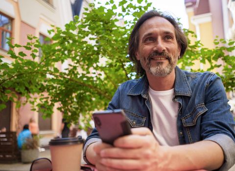 Middle aged man looking away sitting at cafe or street restaurant while working outdoors using digital tablet. Handsome mature freelancer man enjoying coffee outdoors while having group chat.