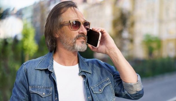Mature bearded man talking on the phone standing outdoors in urban old city background wearing denim jeans shirt. Mature business man answering phone call standing outdoors.