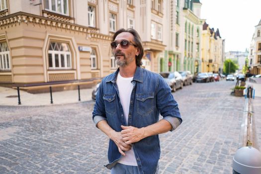 Mature bearded man use walking on urban streets wearing sunglasses and denim jeans shirt. Happy middle aged grey haired business man on old town streets. Travel concept.