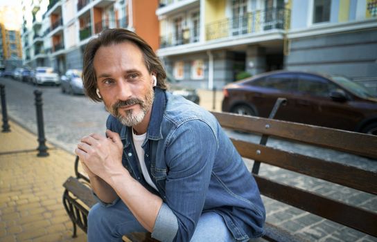 Handsome middle aged man with grey beard spend time talking on the phone or listening music using wireless earphones sitting on the bench in urban city, wearing blue jeans shirt.