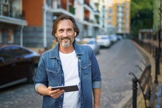 Middle aged grey haired freelancer man hold digital tablet in hands having a call while standing on urban streets. Mature man talking or listening music use wireless earphones travel old town.