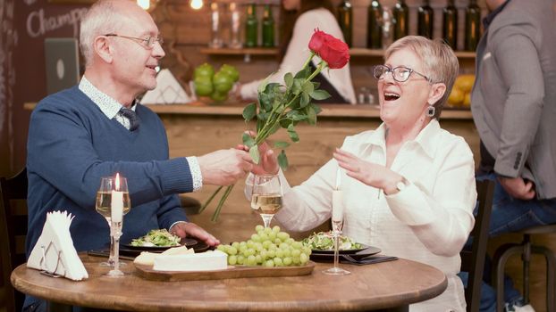 Happy old couple with roses during mealtime in a restaurant. Retirement gift. Happy old woman. Happy senior man.