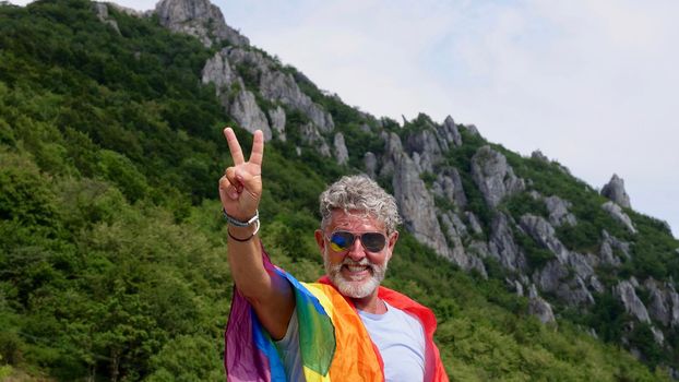 Portrait of a gray-haired senior elderly Caucasian man bisexuality with a beard and sunglasses with a rainbow LGBTQIA peace flag in mountains. Celebrates Pride Month, Rainbow Flag Day, gay parade