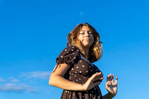 an attractive Caucasian girl with long hair in a fashionable short brown dress against a blue sky in the rays of the setting sun. Stylish teenage girl. Attractive young lady looks at the camera