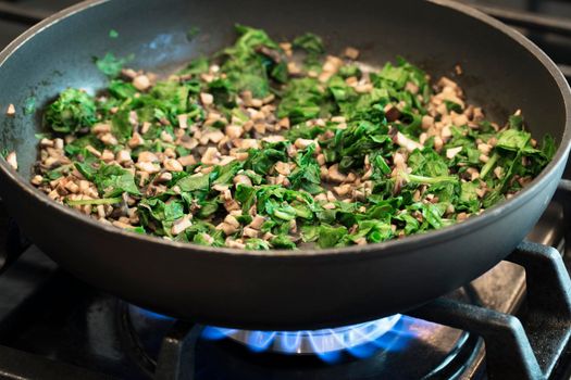 Chopped mushrooms and spinach sautéing in pan on gas burner.