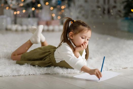 Cute little girl in cozy christmas interior writes letter to Santa against the background of Christmas tree.