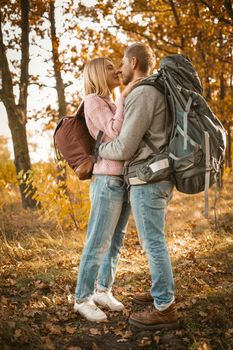 Couple of tourists in love in the autumn forest. Young man and woman kisses and hugs while standing against the backdrop of colorful autumn nature backlit by sunlight. Full-length portrait.