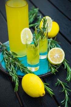Traditional italian lemon alcohol drink limoncello with pieces of lemon and rosemary herb on dark wooden table
