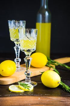 Traditional italian lemon alcohol drink limoncello with pieces of lemon and rosemary herb on dark wooden table