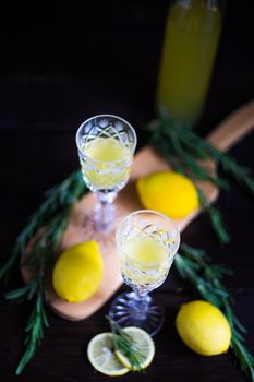 Traditional italian lemon alcohol drink limoncello with pieces of lemon and rosemary herb on dark wooden table