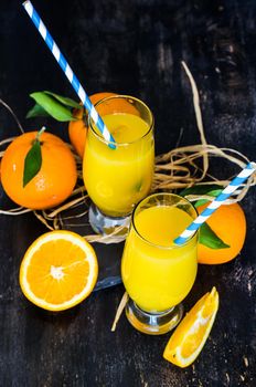 Glass of orange juice with straw and ripe orange fruits with green leaves on dark rustic wooden table