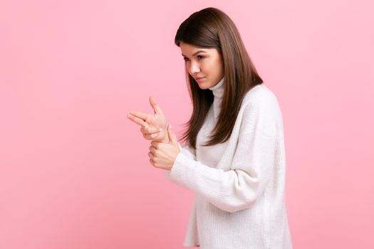 Dangerous criminal woman pointing finger gun, threatening to kill, shooting with weapon gesture, wearing white casual style sweater. Indoor studio shot isolated on pink background.