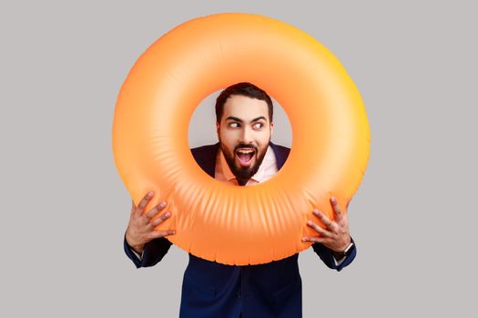 Bearded man holding rubber ring over her neck and looking away with surprised expression and opened mouth, wearing official style suit. Indoor studio shot isolated on gray background.