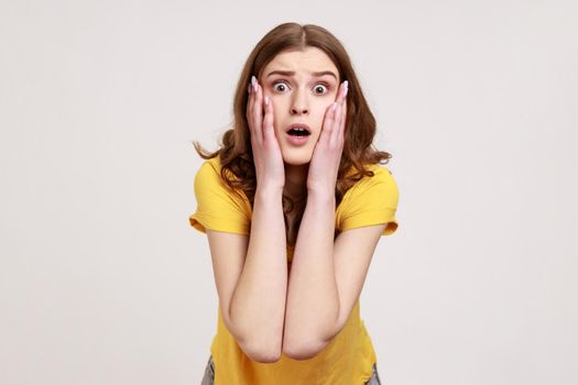 Astonished teenager girl with brown in casual yellow T-shirt standing with mouth open in surprise, has shocked expression, keeps palms on cheeks. Indoor studio shot isolated on gray background.