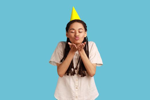 Charming romantic woman party cone hat blowing air kissing over palms hands, sending love, congratulating and celebrating birthday, wearing white shirt. Indoor studio shot isolated on blue background.