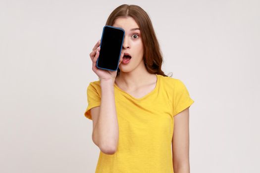 Portrait of excited shocked teenager girl wearing yellow T-shirt covering half face with cell phone and looking at camera with open mouth and big eyes. Indoor studio shot isolated on gray background.