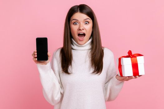 Excited surprised woman holding wrapped present box and cell phone with empty screen for promotion, wearing white casual style sweater. Indoor studio shot isolated on pink background.
