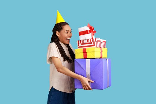 Side view of amazed woman with black dreadlocks, posing in party cone, holding stack of presents, happy birthday, wearing white shirt. Indoor studio shot isolated on blue background.