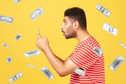Profile side view of money rain and rich surprised shocked man man looking at smartphone screen with shocked face and can not believe. Indoor studio shot isolated on yellow background
