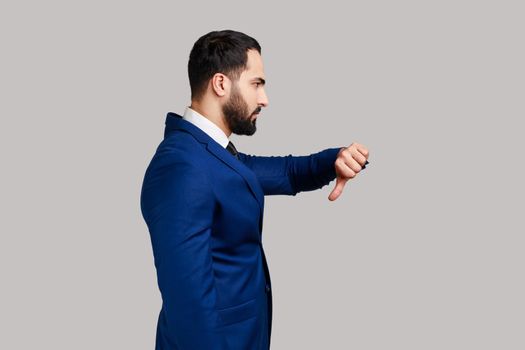 Side view of bearded man criticizing bad quality with thumbs down displeased grimace, showing dislike gesture, expressing disapproval. Indoor studio shot isolated on gray background.