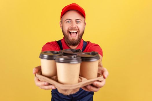 Delivery service. Friendly happy courier man in blue overalls offering coffee, giving drinks in disposable cups, looking at camera with excited look. Indoor studio shot isolated on yellow background.