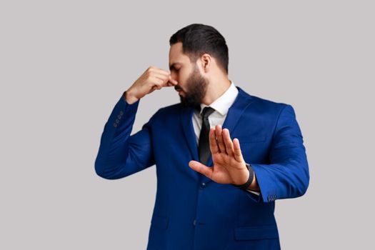 Bearded man grabbing nose, grimacing in disgust and showing stop gesture, expressing repulsion to stink, fart gases, wearing official style suit. Indoor studio shot isolated on gray background.
