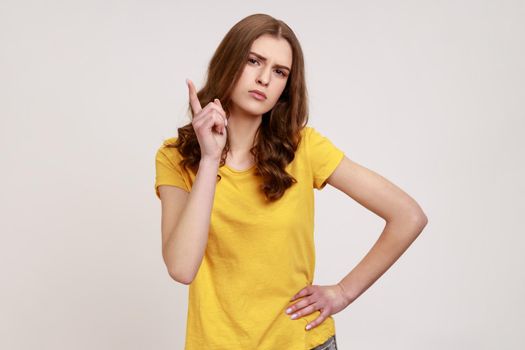 You better do what I say. Portrait of young female wearing yellow casual T-shirt looking at camera with serious face and shows finger warning gesture. Indoor studio shot isolated on gray background.