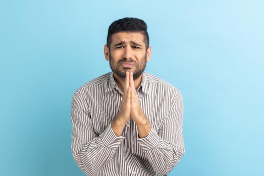 Portrait of businessman keeps palms pressed together, pleading angel, has innocent expressions, asks for apologize, wearing striped shirt. Indoor studio shot isolated on blue background.