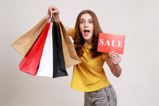 Overjoyed excited teen girl with wavy hair in yellow t-shirt holding sale card and shopping bags, enjoying discounts doing shopping, keeps mouth open. Indoor studio shot isolated on gray background.