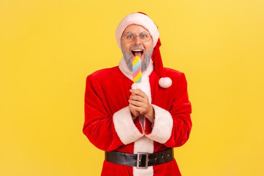 Excited elderly man with gray beard wearing santa claus costume holding and biting multicolored ice cream, celebrating winter holiday. Indoor studio shot isolated on yellow background.