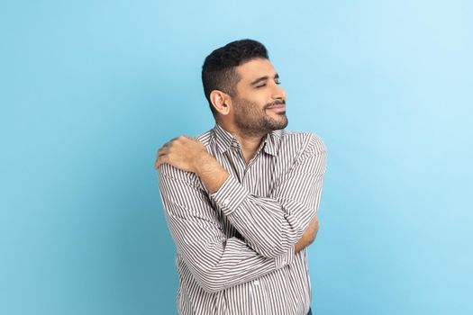 Portrait of happy handsome businessman with beard standing and hugging himself with toothy smile enjoying, positive self-esteem, wearing striped shirt. Indoor studio shot isolated on blue background.