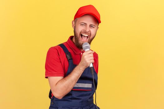 Portrait of happy excited worker man standing with microphone in hands and singing at corporate party, having fun at work with colleagues. Indoor studio shot isolated on yellow background.