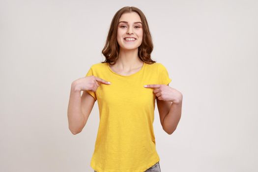 Portrait of charming happy teenager girl in yellow T-shirt smiling and pointing at herself, being proud of own success, boasting of achievement. Indoor studio shot isolated on gray background.