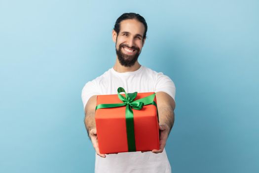 Portrait of handsome delighted man with beard wearing white T-shirt giving wrapped gift box and smiling at camera, happy holiday. Indoor studio shot isolated on blue background.