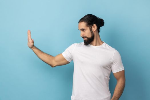 Side view of man with beard wearing white T-shirt showing stop gesture with palm of hand, rejecting stress and problems, no discrimination. Indoor studio shot isolated on blue background.