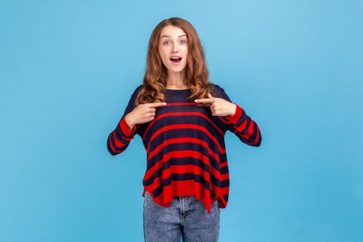 Wow, this is me. Surprised woman wearing striped casual style sweater pointing herself, feeling proud and ambitious, shocked by huge success. Indoor studio shot isolated on blue background.