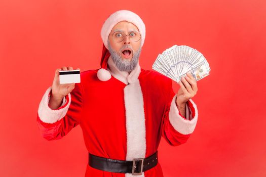 Portrait of shocked elderly man with gray beard wearing santa claus costume holding fan of dollars and credit card, astonished of big cashback. Indoor studio shot isolated on red background.