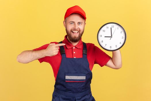 Portrait of delighted man worker standing and pointing at big wall clock in his hands, looking at camera, wearing overalls and red cap. Indoor studio shot isolated on yellow background.