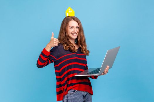 Satisfied woman wearing striped casual style sweater, holding laptop showing thumbs up, looking at camera, real estate service, online renting. Indoor studio shot isolated on blue background.