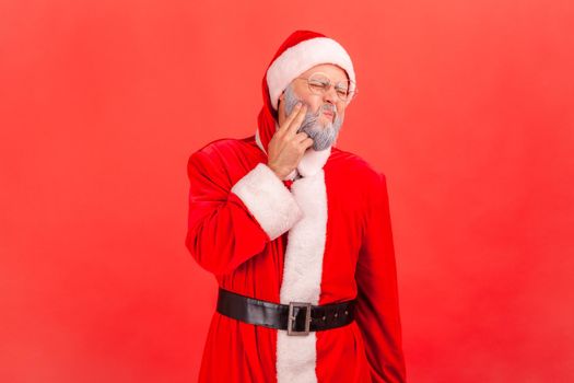 Portrait of sick elderly man with gray beard wearing santa claus costume standing and touching his cheek, suffering from terrible toothache. Indoor studio shot isolated on red background.